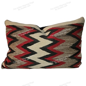 Puede incluir: Cojín decorativo rectangular con un patrón en zigzag en rojo, negro, beige y marrón. Tiene un borde crema y una parte trasera negra. El texto "The Indian Weaving Rugs By Shaifali Jain" está impreso en el cojín.