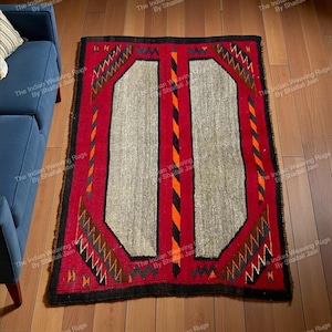 Può includere: Un tappeto rettangolare con bordo rosso e un design centrale con due pannelli grigio chiaro. Il tappeto presenta motivi geometrici neri, arancioni e marroni. Il testo "The Indian Weaving Rugs By Shaifali Jain" è visibile.