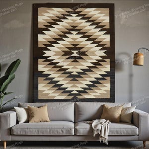 Può includere: Un grande tappeto rettangolare con un motivo geometrico di triangoli nei toni del marrone, beige e nero. Il tappeto è esposto su una parete sopra un divano grigio con cuscini e una coperta. Il tappeto è un elemento decorativo.