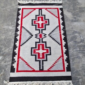 Pode incluir: Tapete retangular tecido com um padrão geométrico. O tapete apresenta um design central em cinza, vermelho e preto, com fundo branco. A borda é preta e o tapete tem franjas nas extremidades curtas. O tapete é feito de material tecido.
