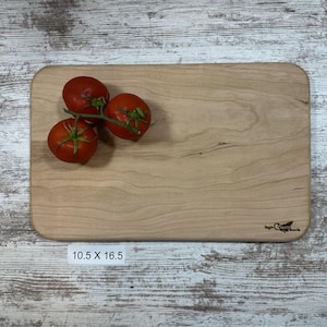 以下が含まれることがあります： 丸みを帯びた角を持つ長方形のメープルウッド製のまな板。まな板は10.5インチ×16.5インチで、自然な木目仕上げ。まな板の上に赤いトマトが3つ置かれている。