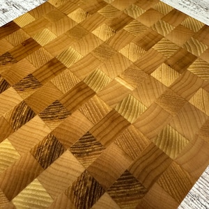 May include: Butcher block cutting board with a woven pattern. The board is made of various shades of light and dark wood, creating a geometric design. The wood grain is visible in each square.