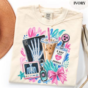 May include: An ivory t-shirt featuring a graphic print of an X-ray of a hand, a chest X-ray monitor, an iced coffee, a teal glove, a pink bow, and an ID badge labeled X RAY TECH with a heart icon, surrounded by colorful abstract brush strokes.