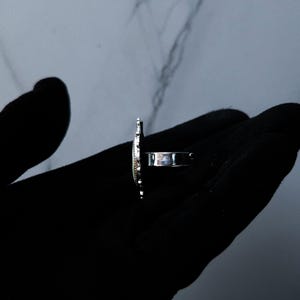 May include: A silver ring with an oval-shaped design. The ring is held in a black gloved hand. The ring's design has a textured surface and a band that wraps around the finger.