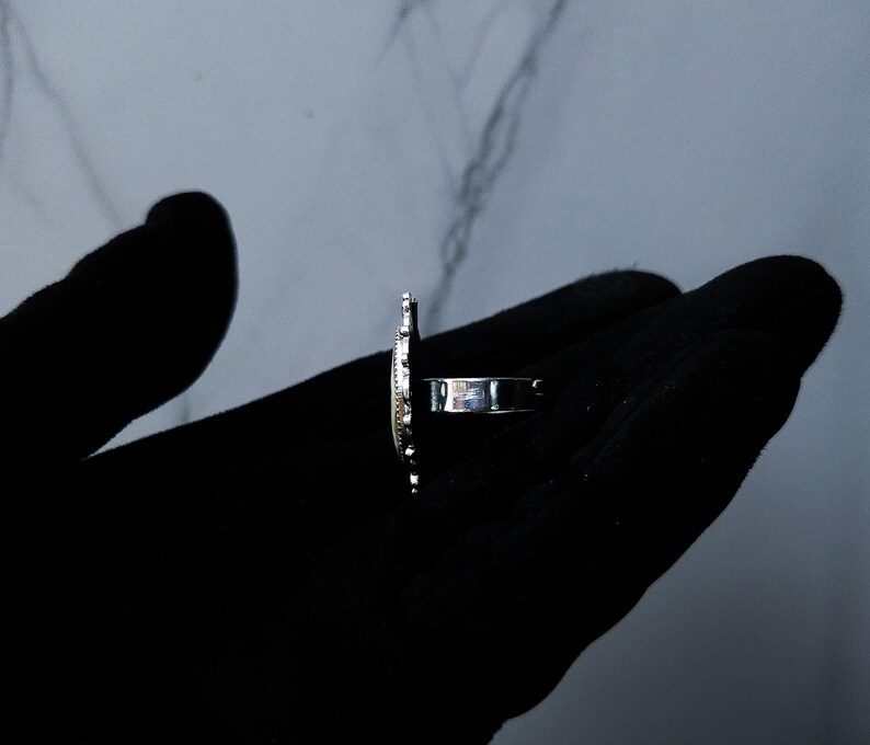 May include: A silver ring with an oval-shaped design. The ring is held in a black gloved hand. The ring's design has a textured surface and a band that wraps around the finger.