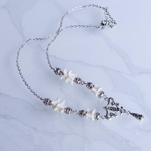 May include: Silver-toned necklace featuring a series of small, white bone-shaped beads and ornate metalwork. The necklace has a delicate chain and a decorative pendant with a teardrop-shaped charm. The necklace is displayed on a white marble surface.