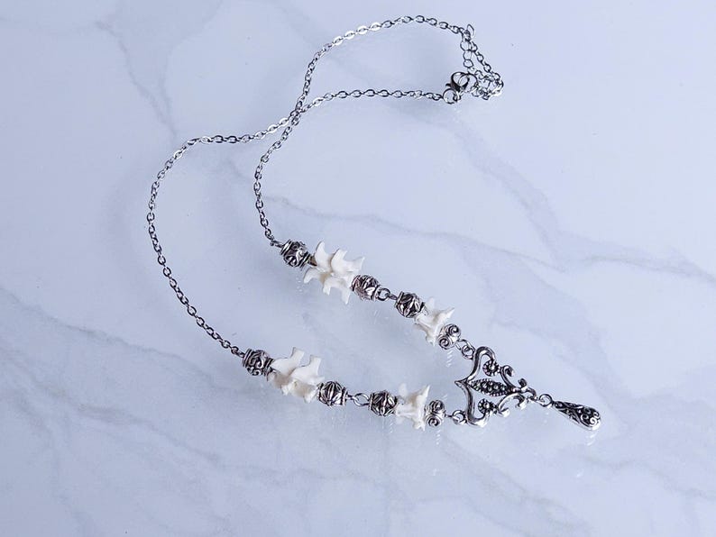 May include: Silver-toned necklace featuring a series of small, white bone-shaped beads and ornate metalwork. The necklace has a delicate chain and a decorative pendant with a teardrop-shaped charm. The necklace is displayed on a white marble surface.