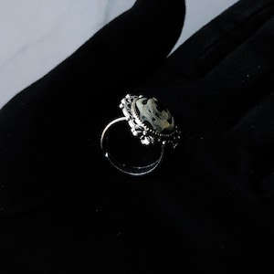 May include: A silver-toned ring with an oval, marbled stone. The stone features a swirling pattern of black, white, and gray. The ring has an ornate, filigree-style setting around the stone, adding a vintage aesthetic.