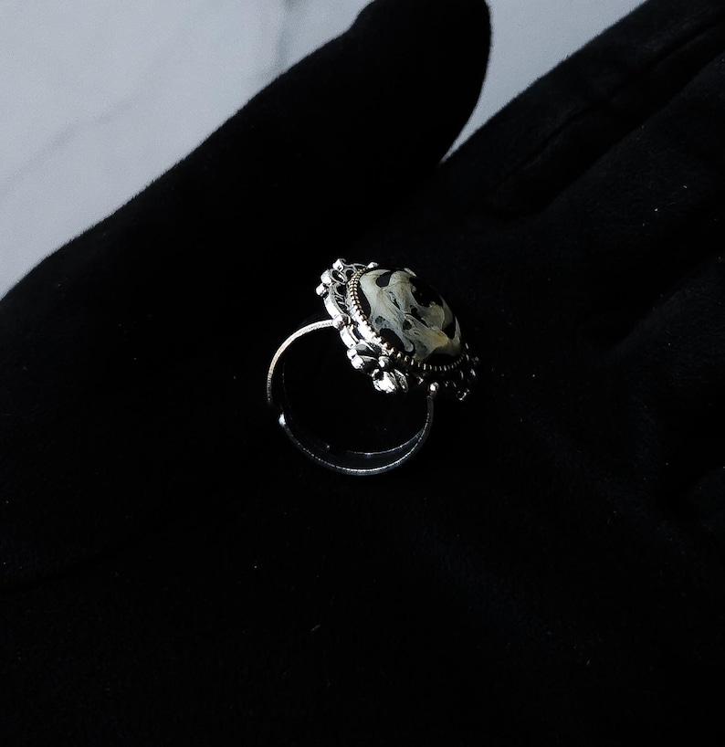May include: A silver-toned ring with an oval, marbled stone. The stone features a swirling pattern of black, white, and gray. The ring has an ornate, filigree-style setting around the stone, adding a vintage aesthetic.