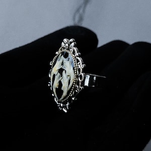 Puede incluir: Anillo plateado con un engaste ovalado y ornamentado. El engaste presenta un fondo negro con un diseño blanco y crema. El anillo se muestra sobre una superficie negra.