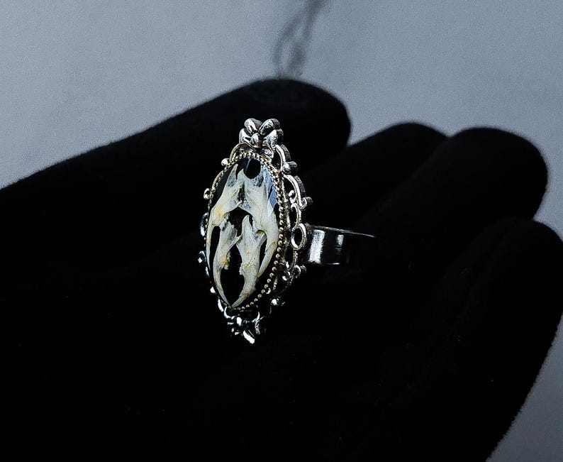 May include: A silver-toned ring with an oval, ornate setting. The setting contains a black background with a white and cream-colored design. The ring is displayed on a black surface.