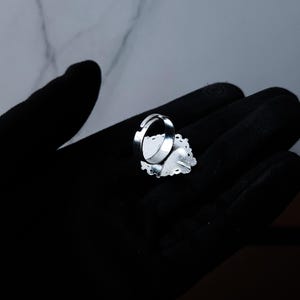 May include: A silver adjustable ring with a decorative oval-shaped face. The face has a white background with a floral design and a heart shape. The ring is set against a black background.