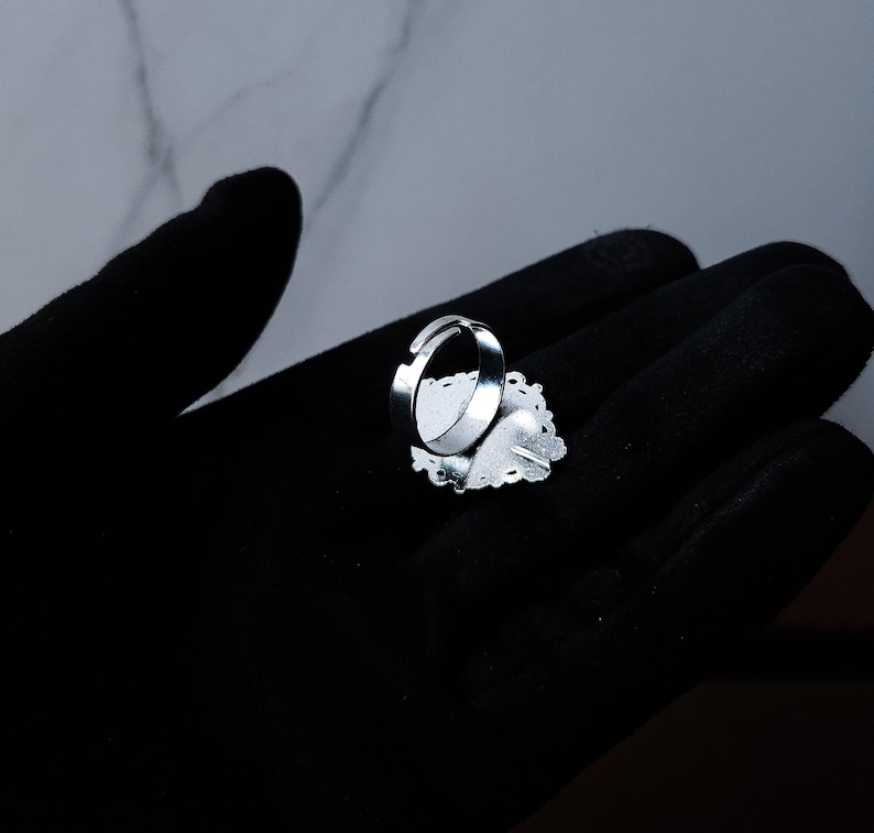 May include: A silver adjustable ring with a decorative oval-shaped face. The face has a white background with a floral design and a heart shape. The ring is set against a black background.