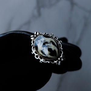 May include: An oval-shaped silver-toned ring with a detailed filigree border. The ring's centerpiece features a dark background with a white and beige organic design, possibly a preserved specimen. The ring is held against a black surface.