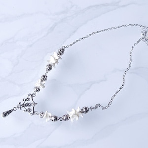 May include: A silver-toned necklace featuring a delicate chain and ornate pendant. The necklace incorporates small, white, bone-shaped beads and decorative metal accents. The design is symmetrical, with a drop pendant at the center.