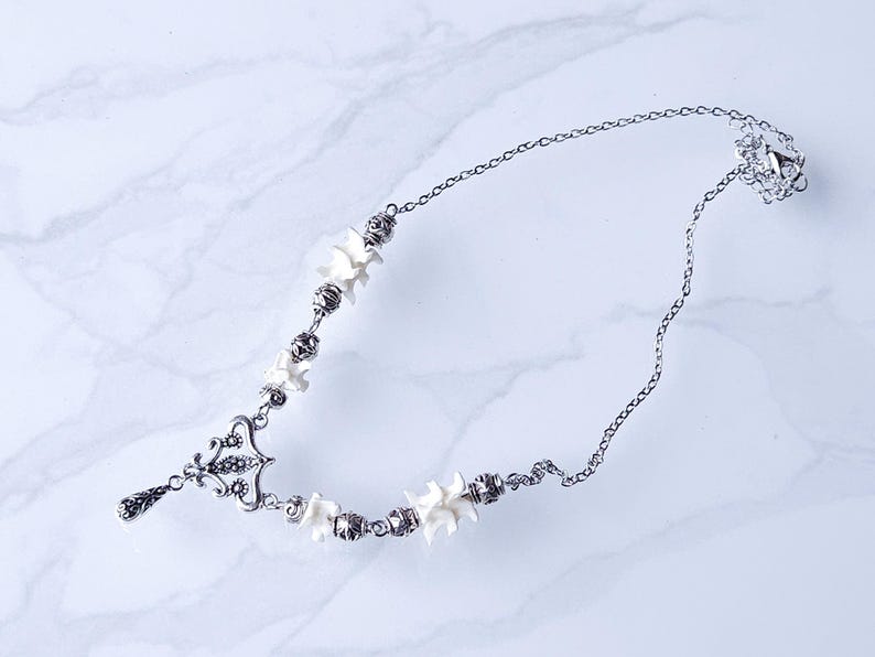 May include: A silver-toned necklace featuring a delicate chain and ornate pendant. The necklace incorporates small, white, bone-shaped beads and decorative metal accents. The design is symmetrical, with a drop pendant at the center.