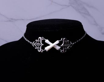 May include: A silver-toned choker necklace featuring ornate filigree designs and a central accent of two crossed white bones. The necklace is displayed on a black mannequin head, showcasing its gothic and alternative style.