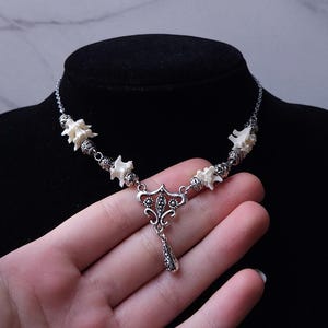 May include: A silver-toned necklace featuring white bone-like elements and decorative metal beads. The pendant has a detailed, ornate design with a teardrop-shaped charm. The necklace is displayed on a black mannequin.