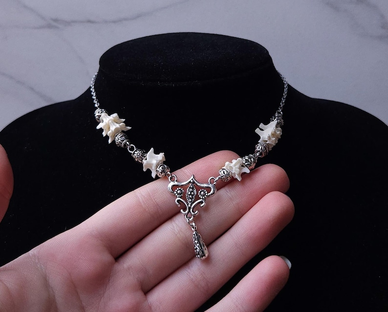 May include: A silver-toned necklace featuring white bone-like elements and decorative metal beads. The pendant has a detailed, ornate design with a teardrop-shaped charm. The necklace is displayed on a black mannequin.