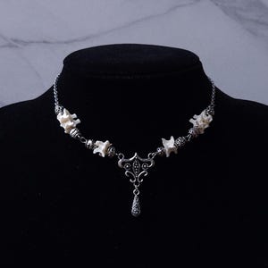 May include: A silver-toned necklace featuring a series of small, white bone segments interspersed with ornate metal beads. The necklace has a decorative pendant with a teardrop-shaped charm. The necklace is displayed on a black mannequin.