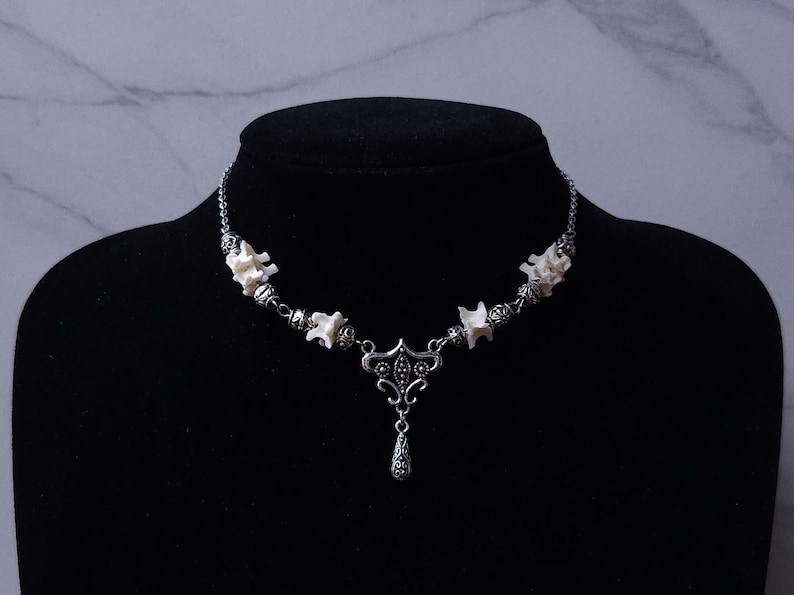 May include: A silver-toned necklace featuring a series of small, white bone segments interspersed with ornate metal beads. The necklace has a decorative pendant with a teardrop-shaped charm. The necklace is displayed on a black mannequin.