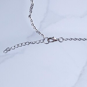 May include: Close-up of a silver-colored necklace chain with a lobster clasp and an extender chain. The chain is on a white and gray marble surface. The necklace is likely for jewelry.