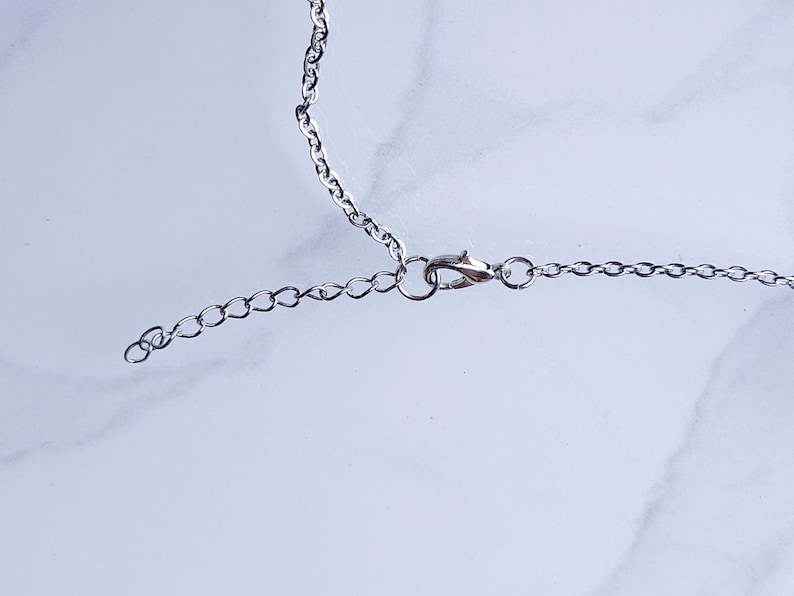 May include: Close-up of a silver-colored necklace chain with a lobster clasp and an extender chain. The chain is on a white and gray marble surface. The necklace is likely for jewelry.