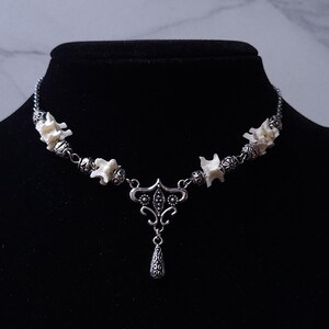 May include: A silver-toned necklace featuring a series of small, off-white bone-like elements interspersed with decorative beads. The necklace has a central pendant with an ornate design and a teardrop-shaped charm. The necklace is displayed on a black mannequin.