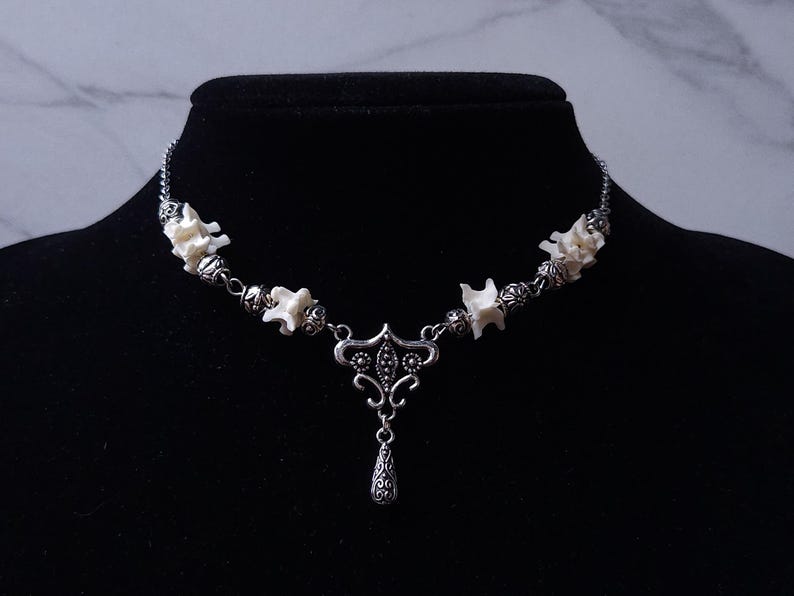 May include: A silver-toned necklace featuring a series of small, off-white bone-like elements interspersed with decorative beads. The necklace has a central pendant with an ornate design and a teardrop-shaped charm. The necklace is displayed on a black mannequin.