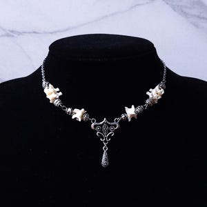 May include: A silver-toned necklace with a gothic aesthetic, featuring bone-shaped beads and a decorative pendant. The necklace is displayed on a black bust against a marble-patterned backdrop.