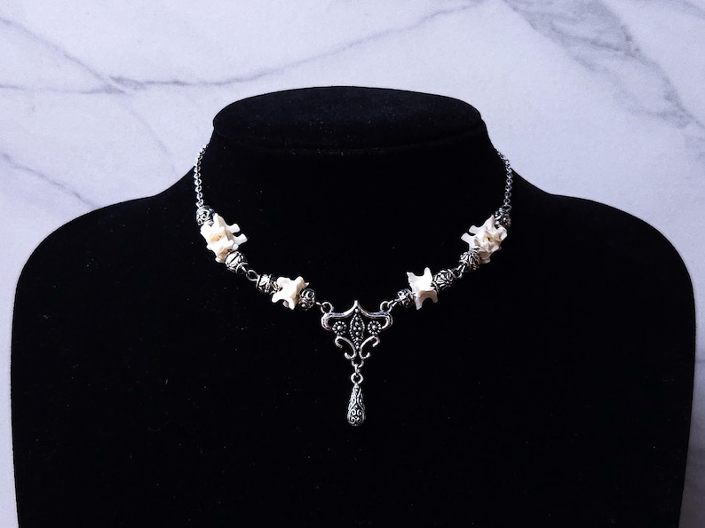 May include: A silver-toned necklace with a gothic aesthetic, featuring bone-shaped beads and a decorative pendant. The necklace is displayed on a black bust against a marble-patterned backdrop.