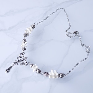 May include: A silver-toned necklace featuring a pendant with an ornate design and a teardrop charm. The necklace incorporates small, white bone-shaped beads and silver beads. The chain is delicate and adjustable, with a lobster clasp closure.