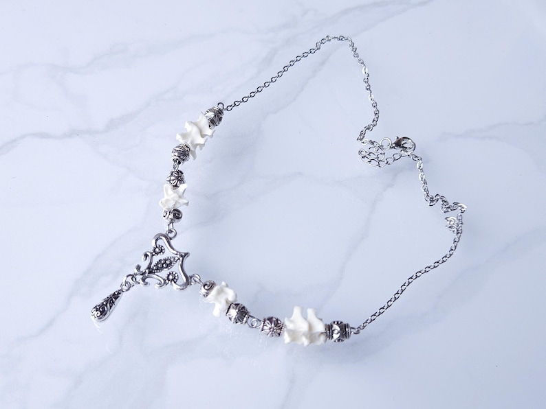 May include: A silver-toned necklace featuring a pendant with an ornate design and a teardrop charm. The necklace incorporates small, white bone-shaped beads and silver beads. The chain is delicate and adjustable, with a lobster clasp closure.