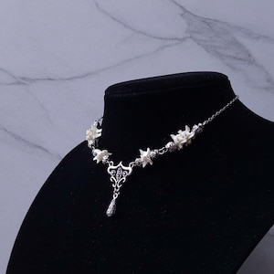 May include: A silver-toned necklace with white floral accents and a teardrop pendant. The necklace is displayed on a black mannequin against a marble-patterned background. The floral elements are evenly spaced along the chain.