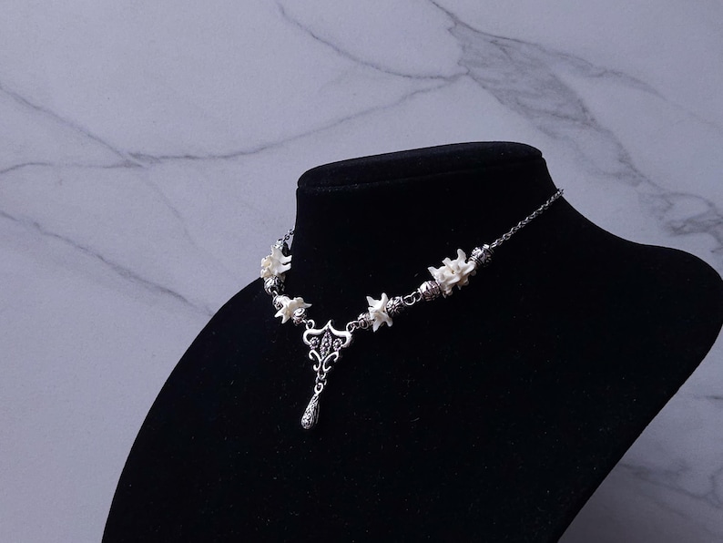 May include: A silver-toned necklace with white floral accents and a teardrop pendant. The necklace is displayed on a black mannequin against a marble-patterned background. The floral elements are evenly spaced along the chain.