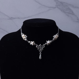 May include: A silver-toned necklace with a unique design, featuring small, white, bone-like elements interspersed with decorative metal beads. A central pendant with a teardrop charm hangs from the necklace, displayed on a black mannequin.