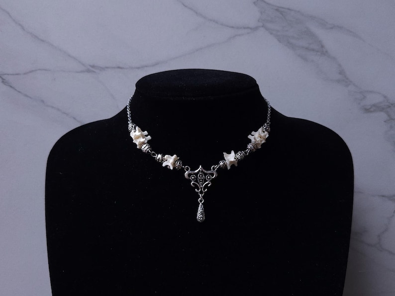 May include: A silver-toned necklace with a unique design, featuring small, white, bone-like elements interspersed with decorative metal beads. A central pendant with a teardrop charm hangs from the necklace, displayed on a black mannequin.