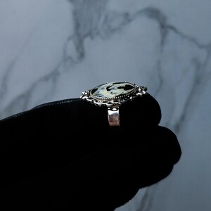 May include: A silver ring with an oval-shaped, black and white patterned stone. The ring has an ornate, filigree-style setting. The ring is shown on a black surface against a marbled background.