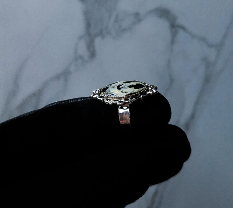 May include: A silver ring with an oval-shaped, black and white patterned stone. The ring has an ornate, filigree-style setting. The ring is shown on a black surface against a marbled background.