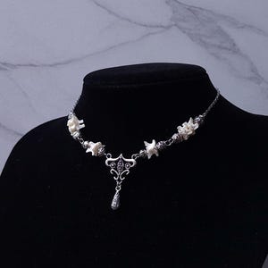 May include: A silver-toned necklace with a pendant and bone-shaped accents. The necklace is displayed on a black mannequin. The pendant has a teardrop shape and intricate detailing. The bone accents are white.