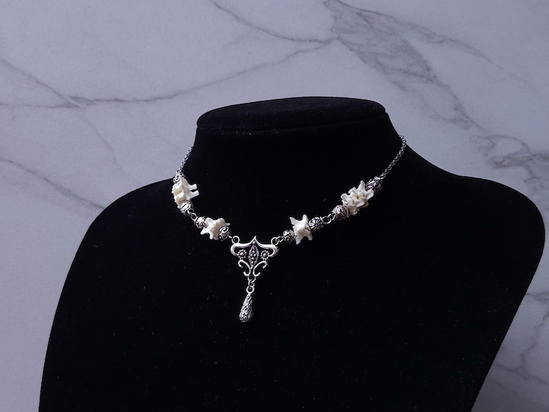 May include: A silver-toned necklace with a pendant and bone-shaped accents. The necklace is displayed on a black mannequin. The pendant has a teardrop shape and intricate detailing. The bone accents are white.