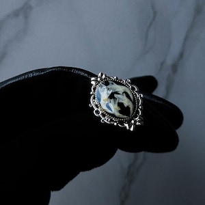 May include: An ornate silver-toned ring with a central oval-shaped design. The design features a black background with white and blue floral elements. The ring is held against a black surface.