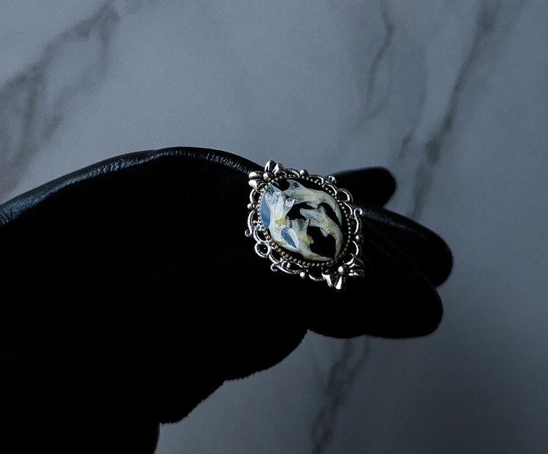 May include: An ornate silver-toned ring with a central oval-shaped design. The design features a black background with white and blue floral elements. The ring is held against a black surface.