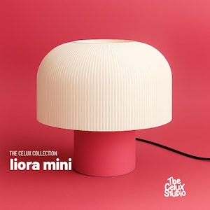 May include: A contemporary table lamp featuring a cream-coloured, ribbed lampshade and a bright pink base. The lamp is labelled "liora mini" from "The Celux Collection" against a red backdrop. A black power cord extends from the base.