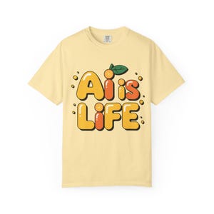 Puede incluir: Camiseta amarilla clara con el texto "Ai is LIFE" en una fuente de burbujas. El texto es amarillo y naranja con un pequeño gráfico de naranja y hoja verde encima de la "is".