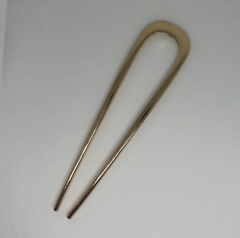 Peut inclure: Une &eacute;pingle &agrave; cheveux dor&eacute;e avec un dessus en forme de U et deux longues broches pointues. L'&eacute;pingle est en m&eacute;tal et a une finition lisse et polie. L'accessoire pour cheveux est con&ccedil;u pour maintenir les cheveux en place.