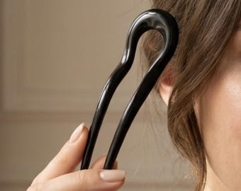 May include: A smooth, glossy black U-shaped hair fork with a curved, minimalist design. The hair accessory features two long, tapered prongs connected by a rounded top, crafted from a solid, reflective material that catches the light.