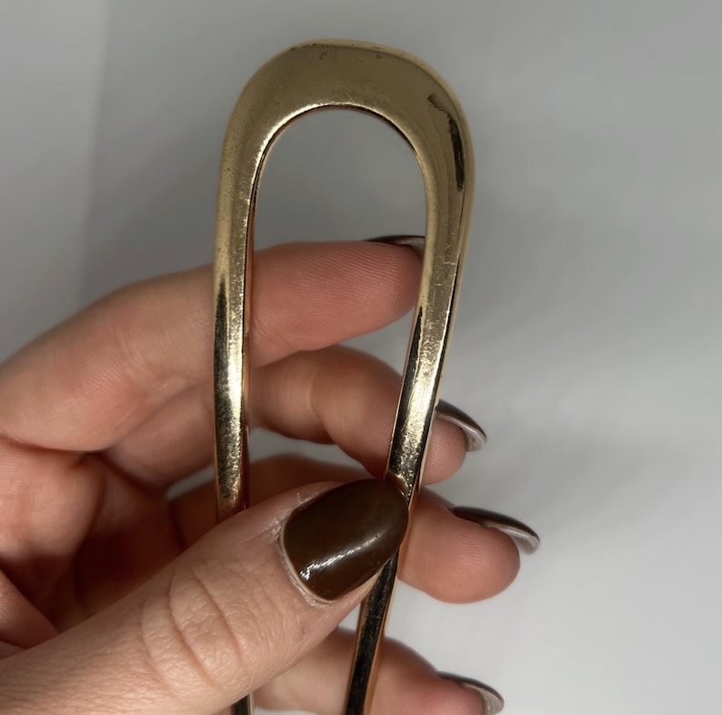 Peut inclure: Une &eacute;pingle &agrave; cheveux dor&eacute;e en forme de U. L'&eacute;pingle est en m&eacute;tal et a une surface lisse et brillante. L'accessoire pour cheveux est tenu dans une main sur fond blanc.