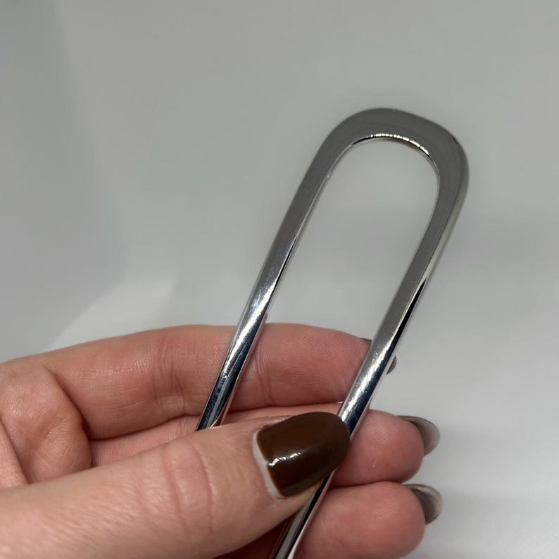 Peut inclure: Une &eacute;pingle &agrave; cheveux argent&eacute;e brillante en forme de U. L'&eacute;pingle est tenue dans une main avec un ongle peint en marron. L'arri&egrave;re-plan est de couleur gris clair uni.
