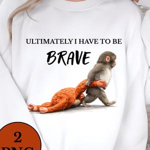 以下が含まれることがあります： 「ULTIMATELY I HAVE TO BE BRAVE」の文字と、ぬいぐるみのおもちゃを引っ張る猿のイラストが描かれた白いスウェットシャツ。左下に「2 PNG」と書かれた茶色の円があります。スウェットシャツは長袖です。